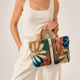 A woman wearing a cream ribbed tank top and white pants holds a woven handbag with a multicolor botanical pattern in blue, beige, pink, and brown. She is photographed against a plain light beige studio background.