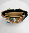 A top-down view of a decorative handbag with a multicolored beaded exterior, gold-toned bamboo-style handles, and a black interior lining with a beige pouch, set against a white background.