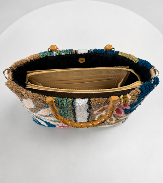 A top-down view of a decorative handbag with a multicolored beaded exterior, gold-toned bamboo-style handles, and a black interior lining with a beige pouch, set against a white background.