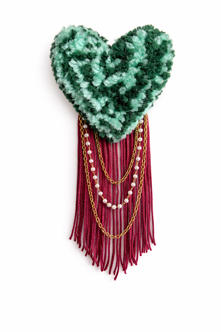 Heart-shaped green knitted piece with red fringe and gold beads on a white background